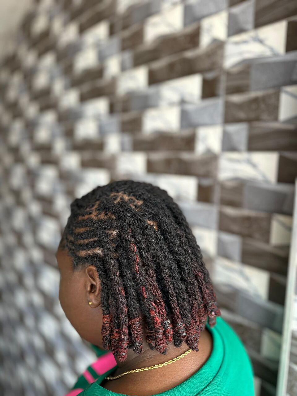 Shoulder-length locs with deep red ends, seen from the side against a tiled wall. The client wears a green top and a gold chain.