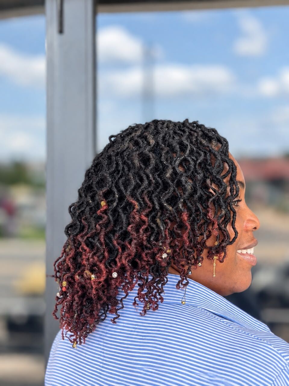 A smiling client in profile with shoulder-length curled locs that fade to burgundy at the ends, decorated with small beads.